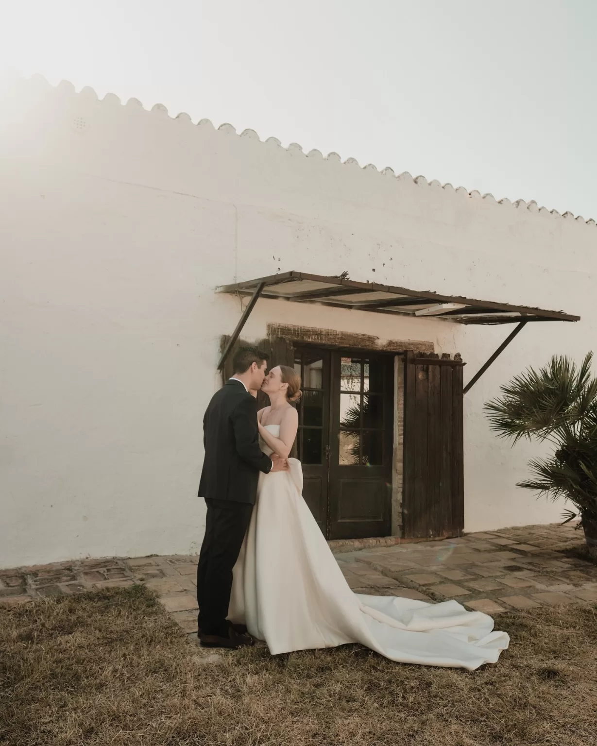 Bride and groom sharing a romantic kiss outside the sunlit white walls of Masia Can Ramonet.