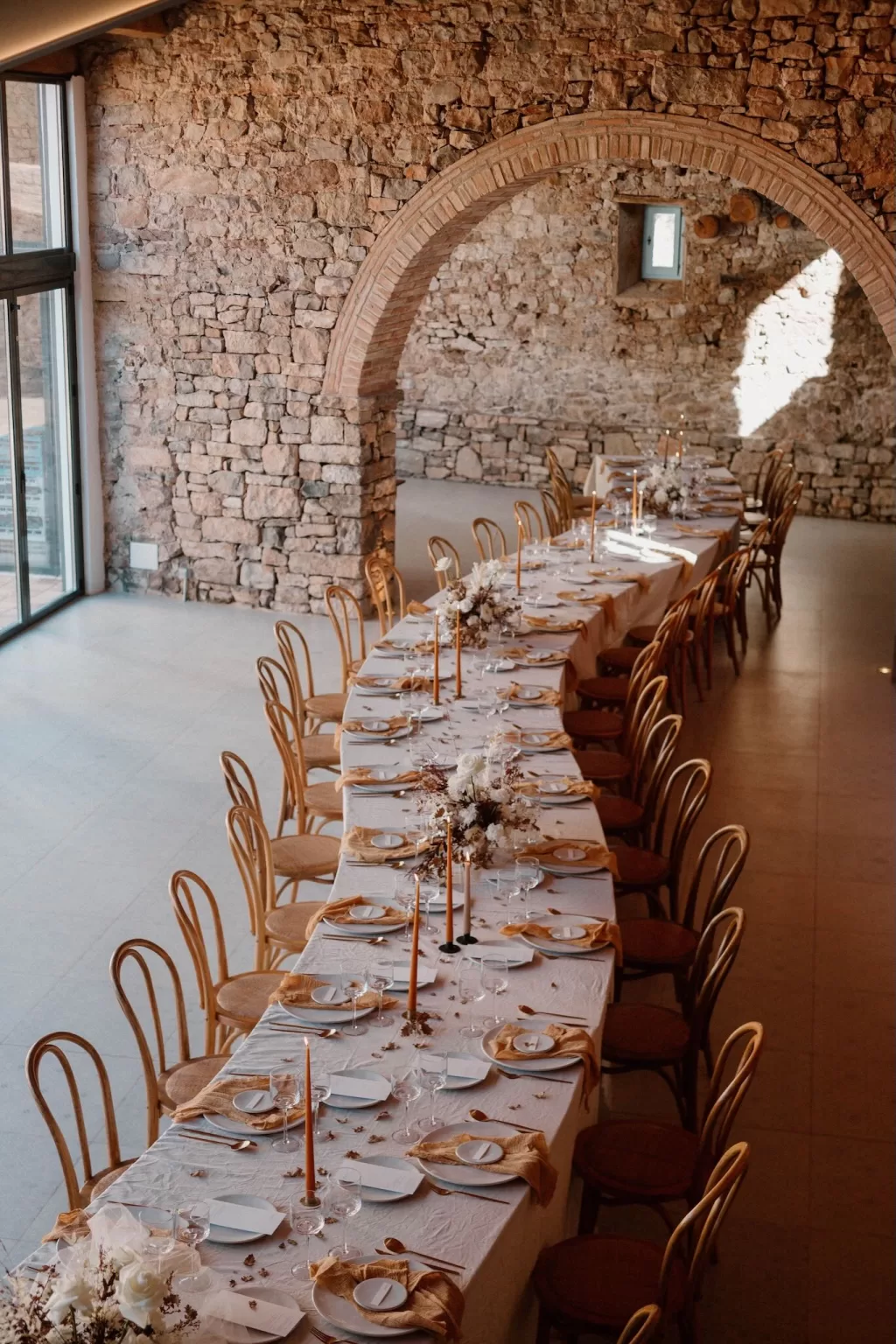 Elegant wedding table setup at This Must Be The Place with curved banquet layout, neutral tones, and soft natural light against stone walls.