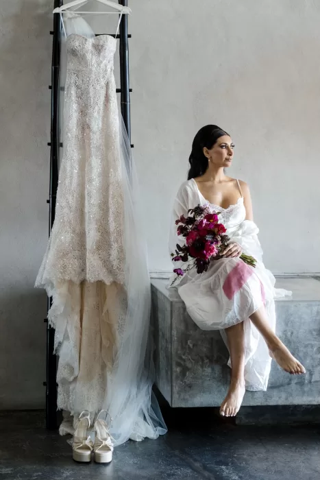 Bride sits beside her lace wedding dress and veil at Areias do Seixo, holding a vibrant bouquet with deep pink tones.