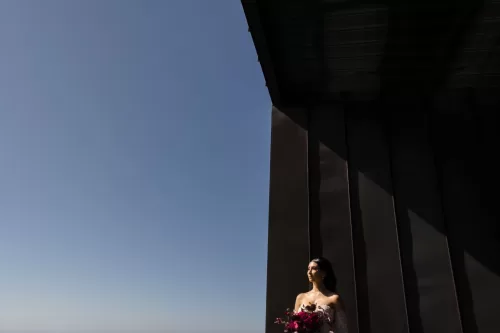 The bride stands in dramatic light against the modern architecture of Areias do Seixo, holding a bright floral bouquet.