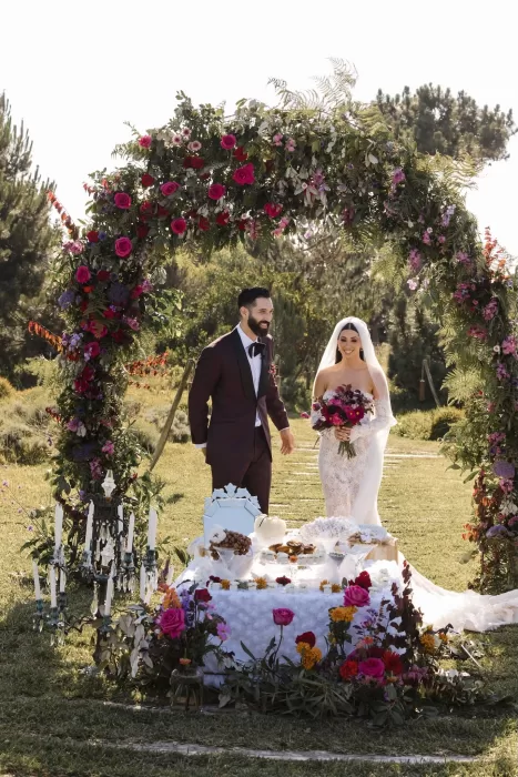 The couple stands beneath a lush floral arch at Areias do Seixo, surrounded by colourful blooms and sunlight.