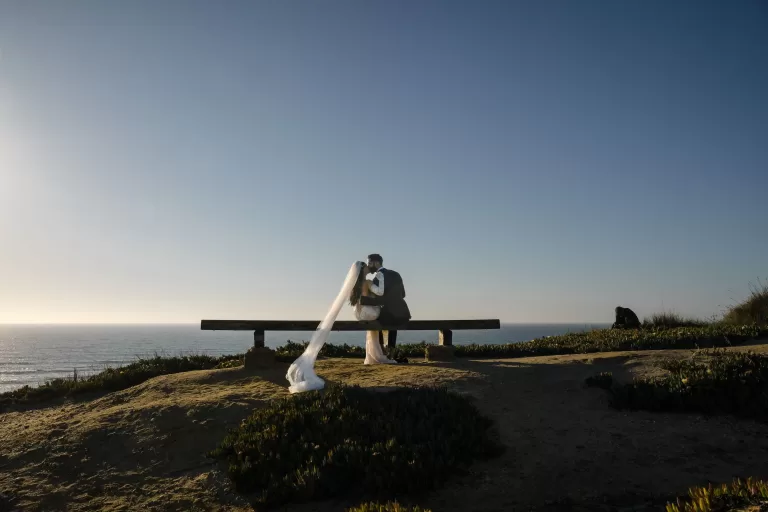 Couple shares a quiet moment on a bench overlooking the ocean at Areias do Seixo, the bride’s veil trailing in the wind.
