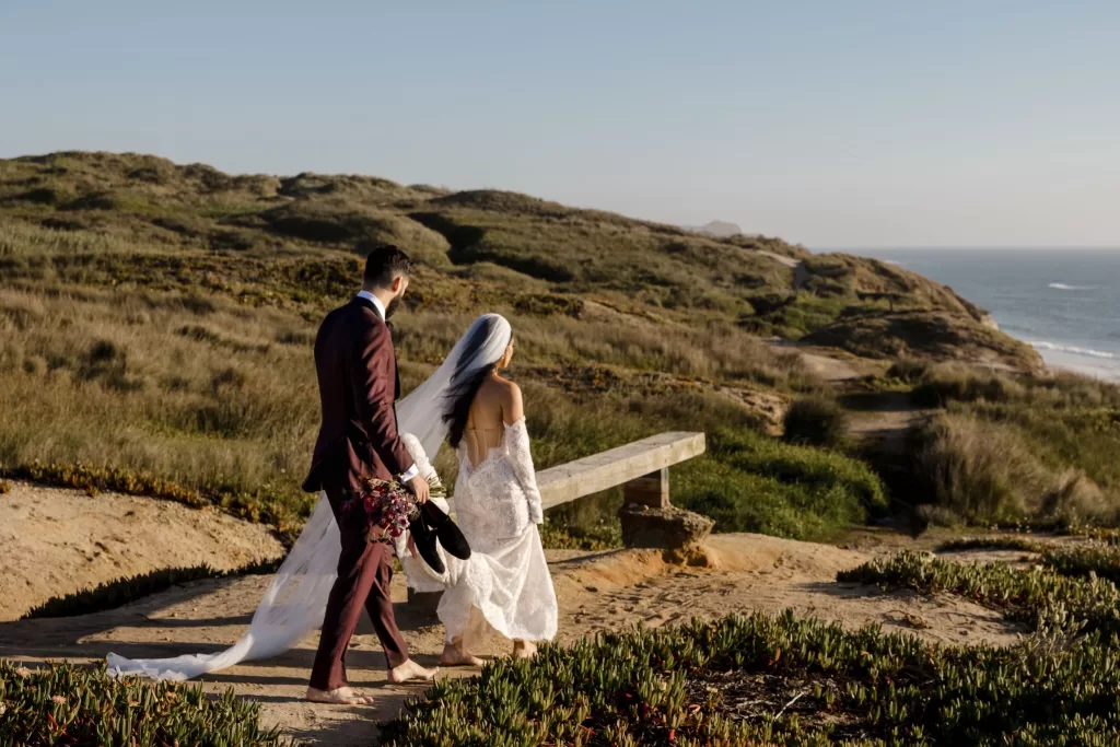 Bride and groom walk along the coastal path near Areias do Seixo, overlooking rolling dunes and the Atlantic Ocean.