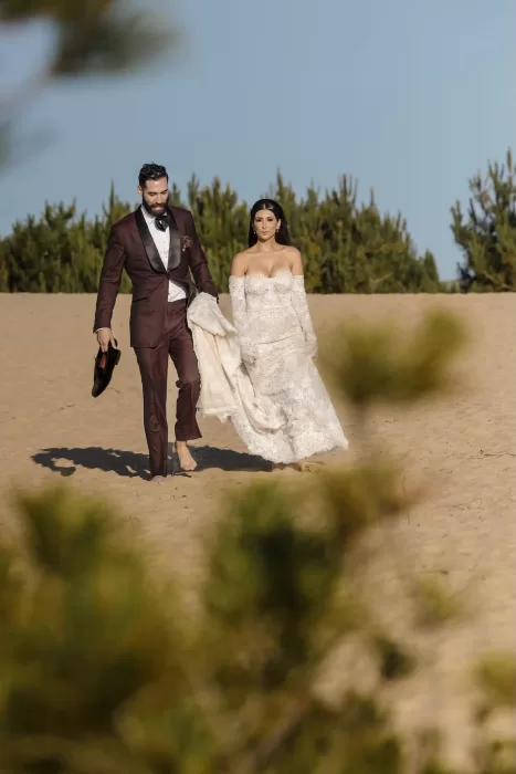Bride and groom walk barefoot across the golden dunes near Areias do Seixo, carrying their wedding attire.