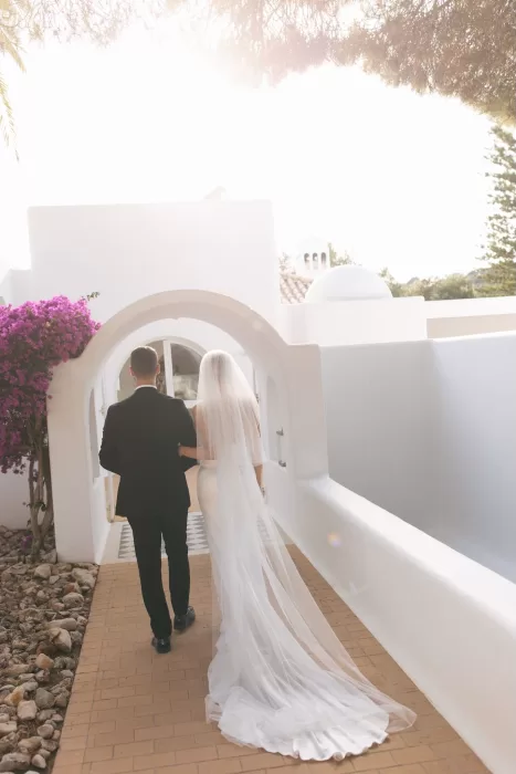 Couple walking hand in hand beneath the white arched walkway at Casa Arte, surrounded by bougainvillea and glowing afternoon sun.