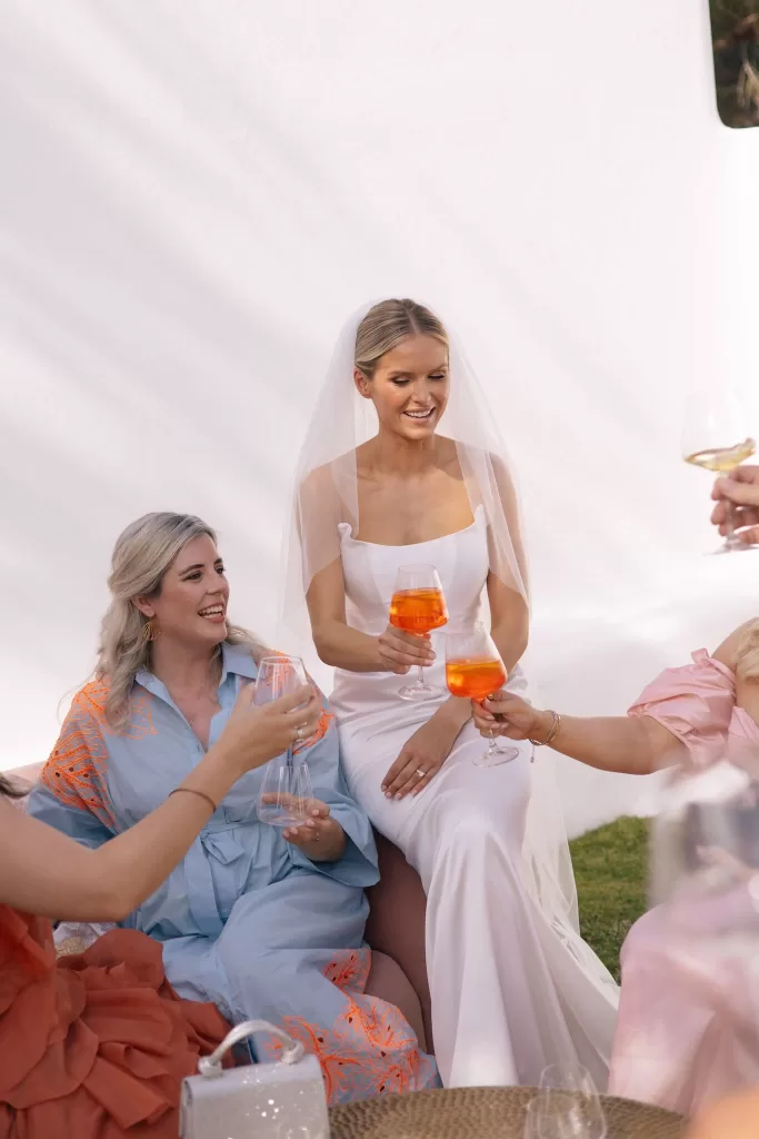 Bride enjoying celebratory drinks with friends at Casa Arte, sitting against the venue’s sunlit white walls.