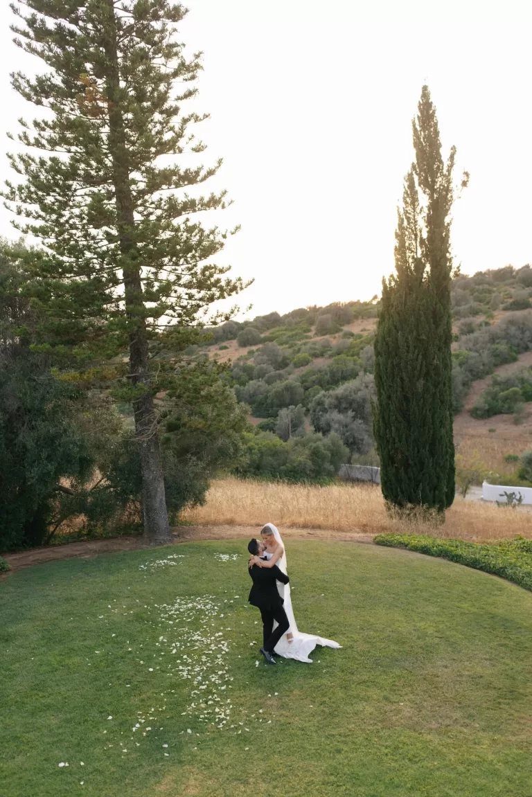 Bride and groom embracing on the lawn at Casa Arte, surrounded by hillside views and tall cypress trees at sunset.