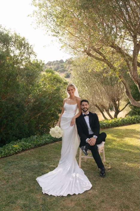 Bride and groom posing beneath sunlit olive trees at Casa Arte, her long satin gown flowing across the grass.