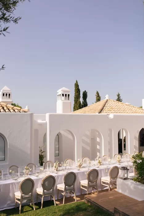 Long outdoor dining table at Casa Arte set with white linens, soft florals, and elegant chairs beneath the crisp white arches of the villa.