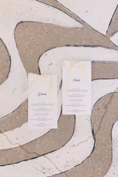 Bride and groom menus tied with soft ivory ribbons, styled as a flatlay on Casa Arte’s textured mosaic floor.