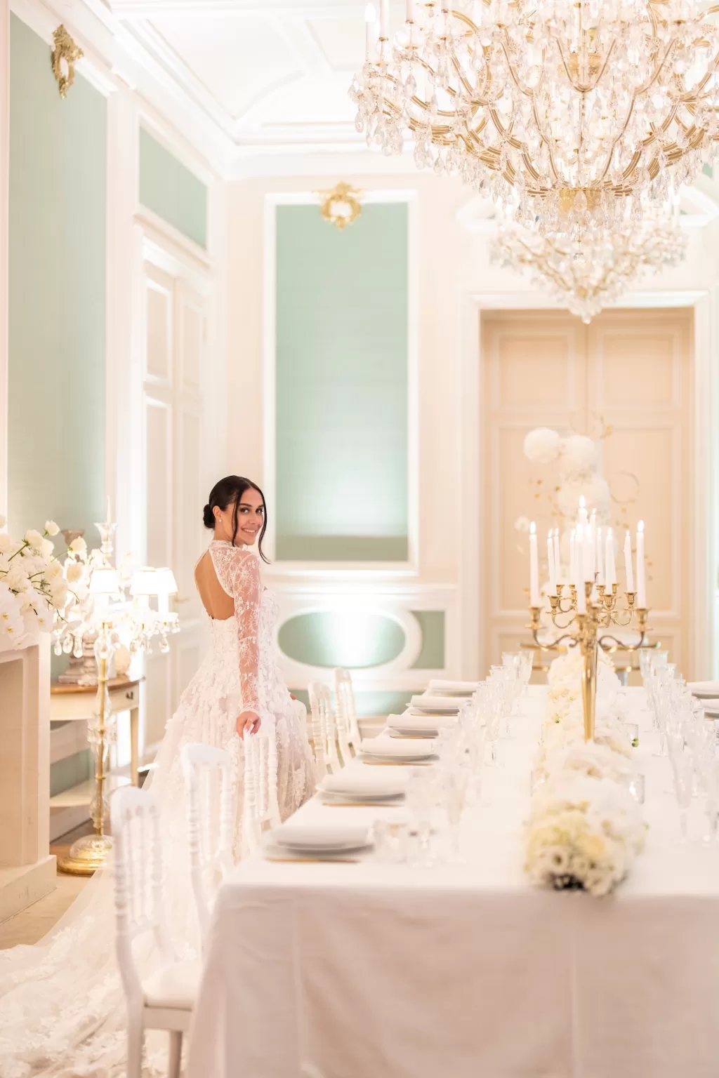 Bride posing beside an elegant white tablescape beneath sparkling chandeliers in Château Bouffémont’s formal dining room.