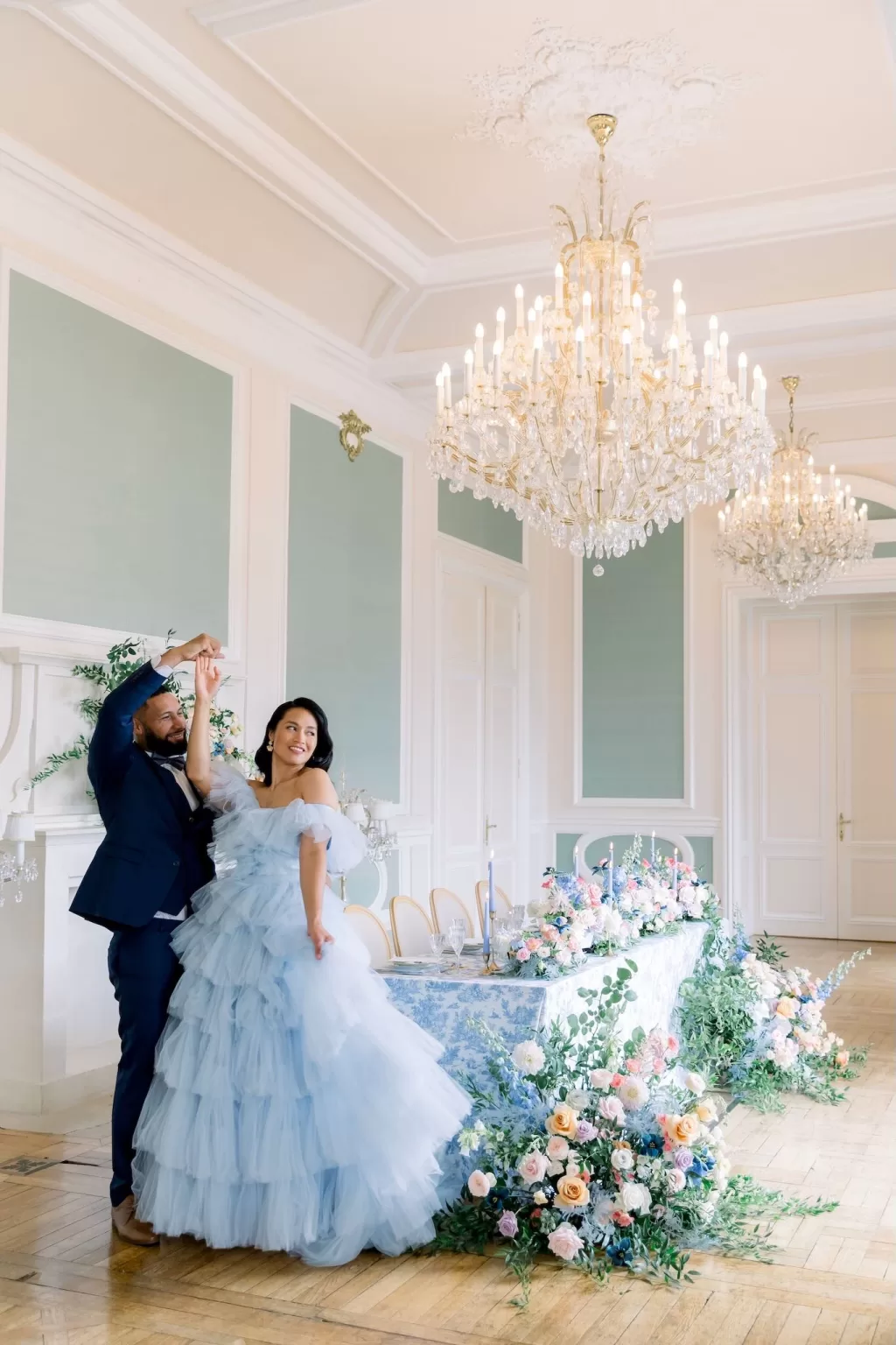 Couple dancing beside an elegant pastel-floral tablescape beneath sparkling chandeliers inside Château Bouffémont.