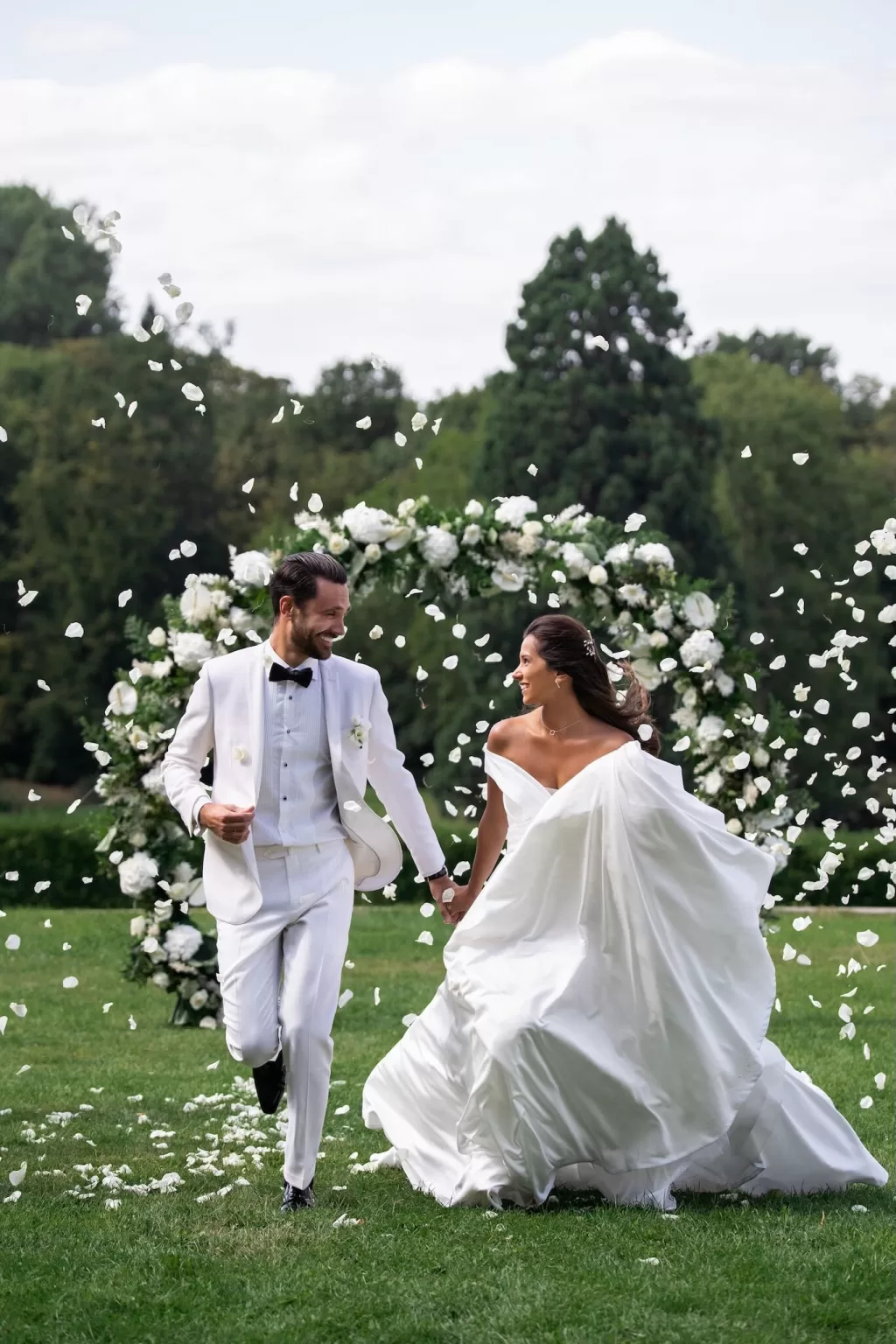 Couple running hand-in-hand across the lawn at Château Bouffémont as white petals fall around them, with a lush floral arch behind.