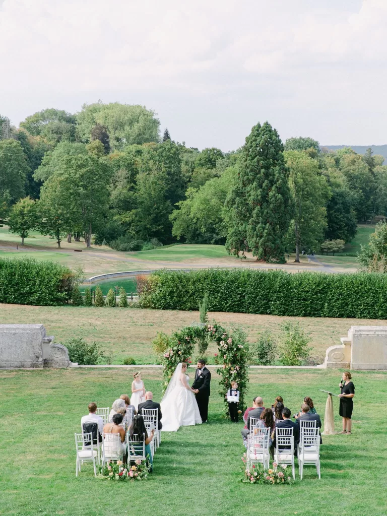 A couple exchanging vows under a lush floral arch on the gardens of Château Bouffémont, framed by rolling greenery.