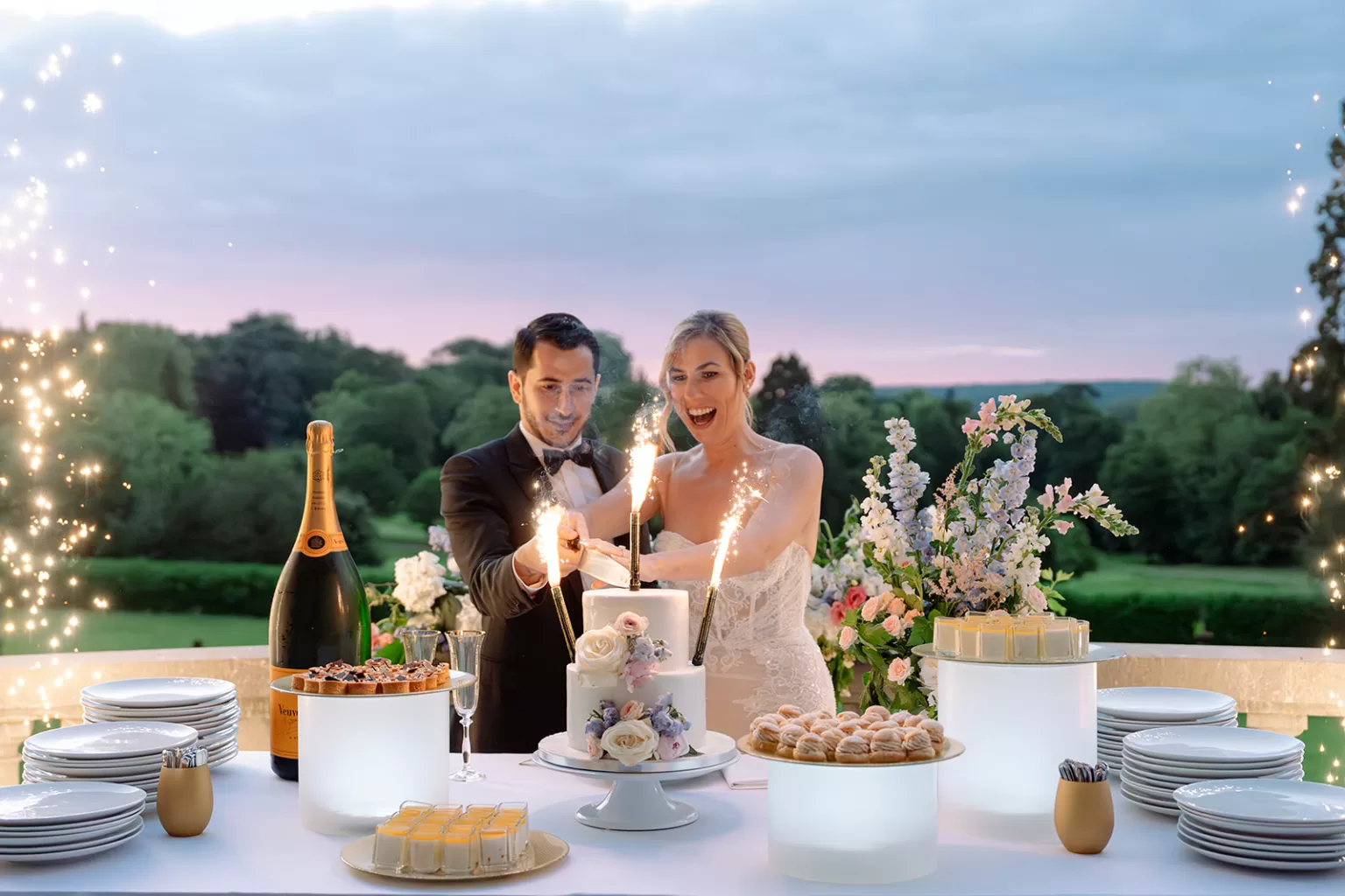 Newlyweds cutting their cake with sparklers glowing around them on the terrace at Château Bouffémont during sunset.
