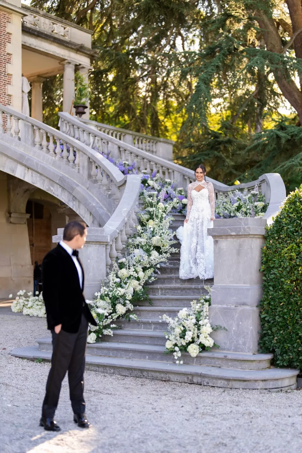 Bride descending the grand outdoor staircase of Château Bouffémont lined with soft white floral arrangements, groom waiting below.
