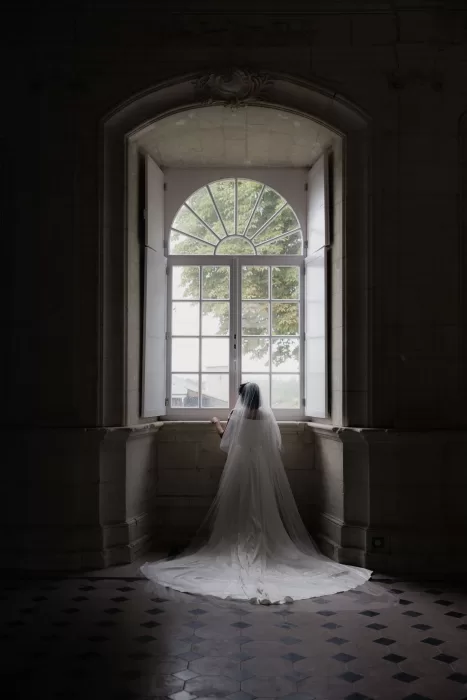 Bride in flowing veil standing before a tall arched window inside Château de Jalesnes, softly lit by daylight.