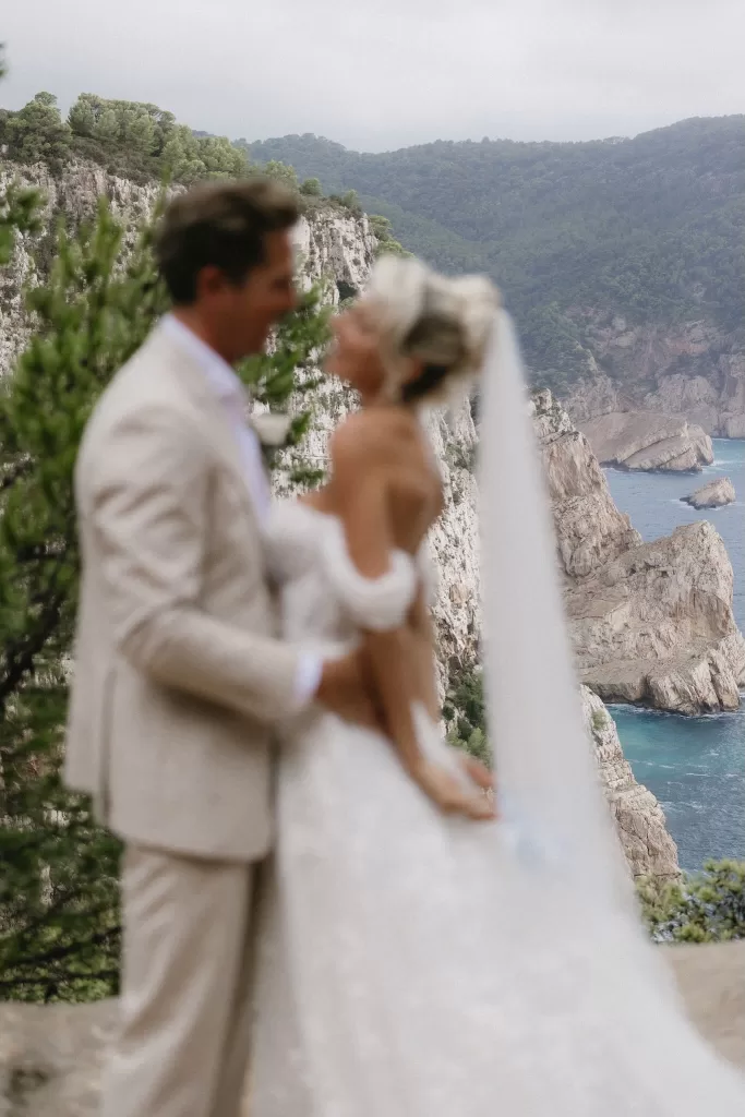 Softly focused image of the couple with the rugged coastline and turquoise waters of Hacienda Na Xamena behind them.