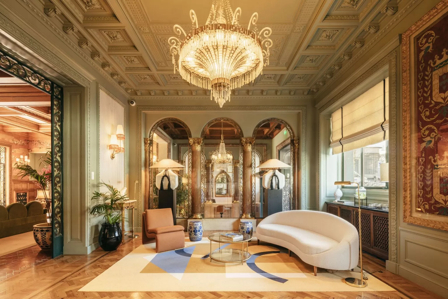 Elegant lounge at Hospes Infante Sagres with crystal chandelier, arched mirrors, and modern curved sofa under ornate ceiling.