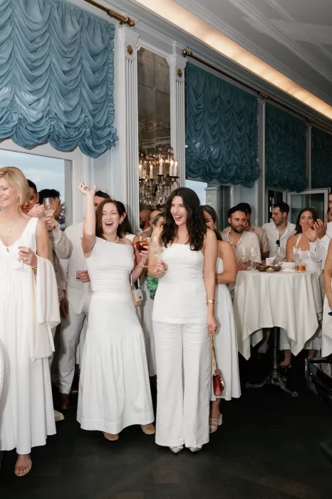 Guests in elegant white attire celebrating at Hotel Château Gütsch, surrounded by chandeliers and blue drapes.