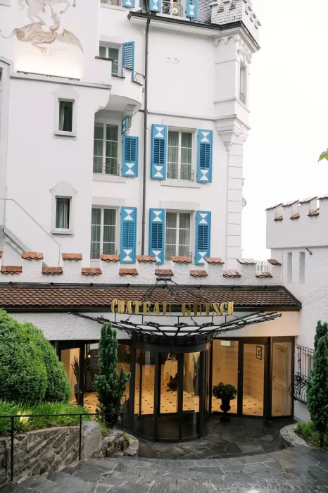 Entrance of Hotel Château Gütsch with blue shutters, tiled roof, and golden signage welcoming guests.