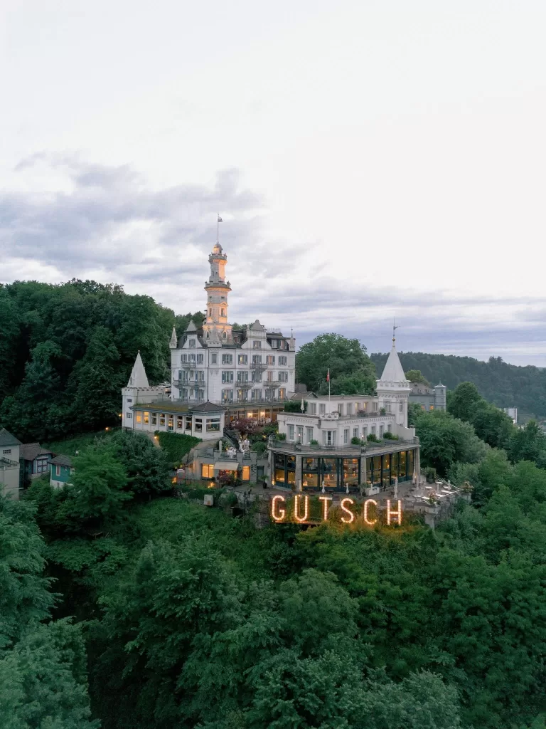 Aerial view of Hotel Château Gütsch perched on a lush hillside in Lucerne, glowing warmly against the evening sky.