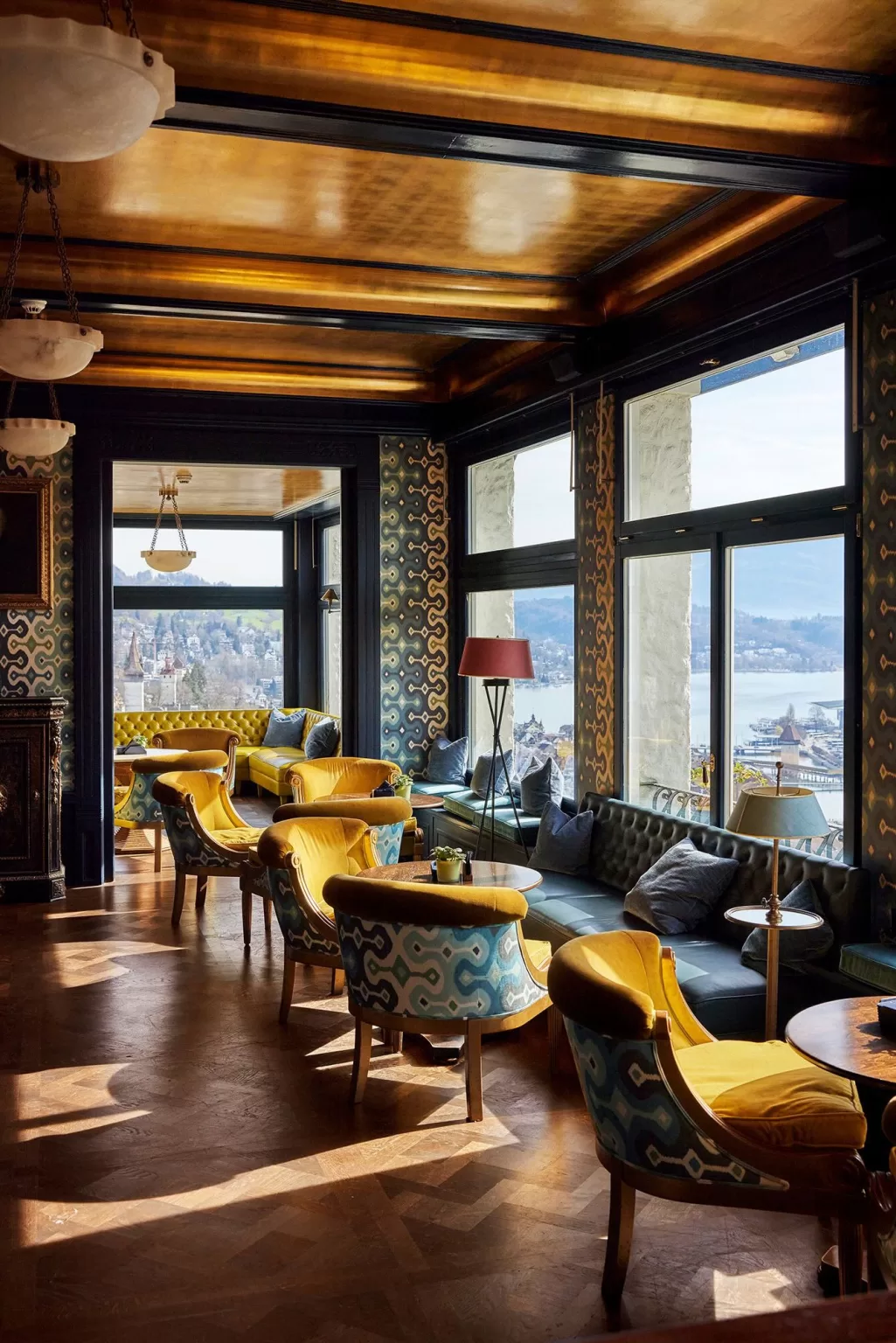 Elegant lounge at Hotel Château Gütsch with patterned walls, mustard velvet chairs, and panoramic views over Lucerne.