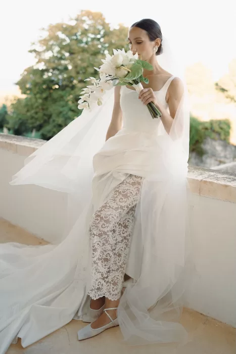 Bride seated outdoors at Masseria Spina, holding a soft white bouquet with lace trousers peeking beneath her gown.