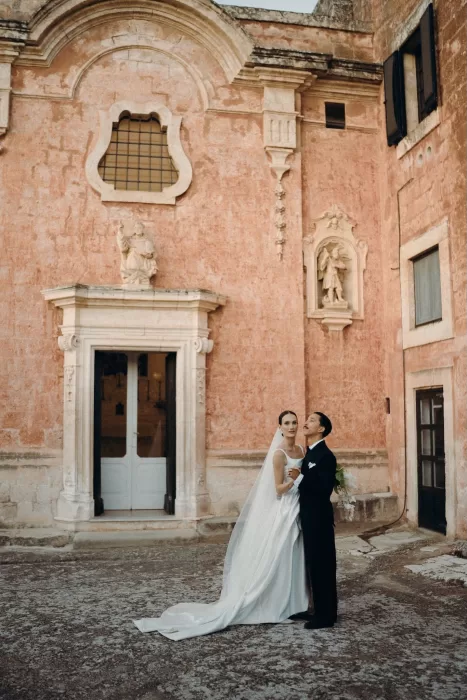 Bride and groom standing in front of the weathered pink façade of Masseria Spina.
