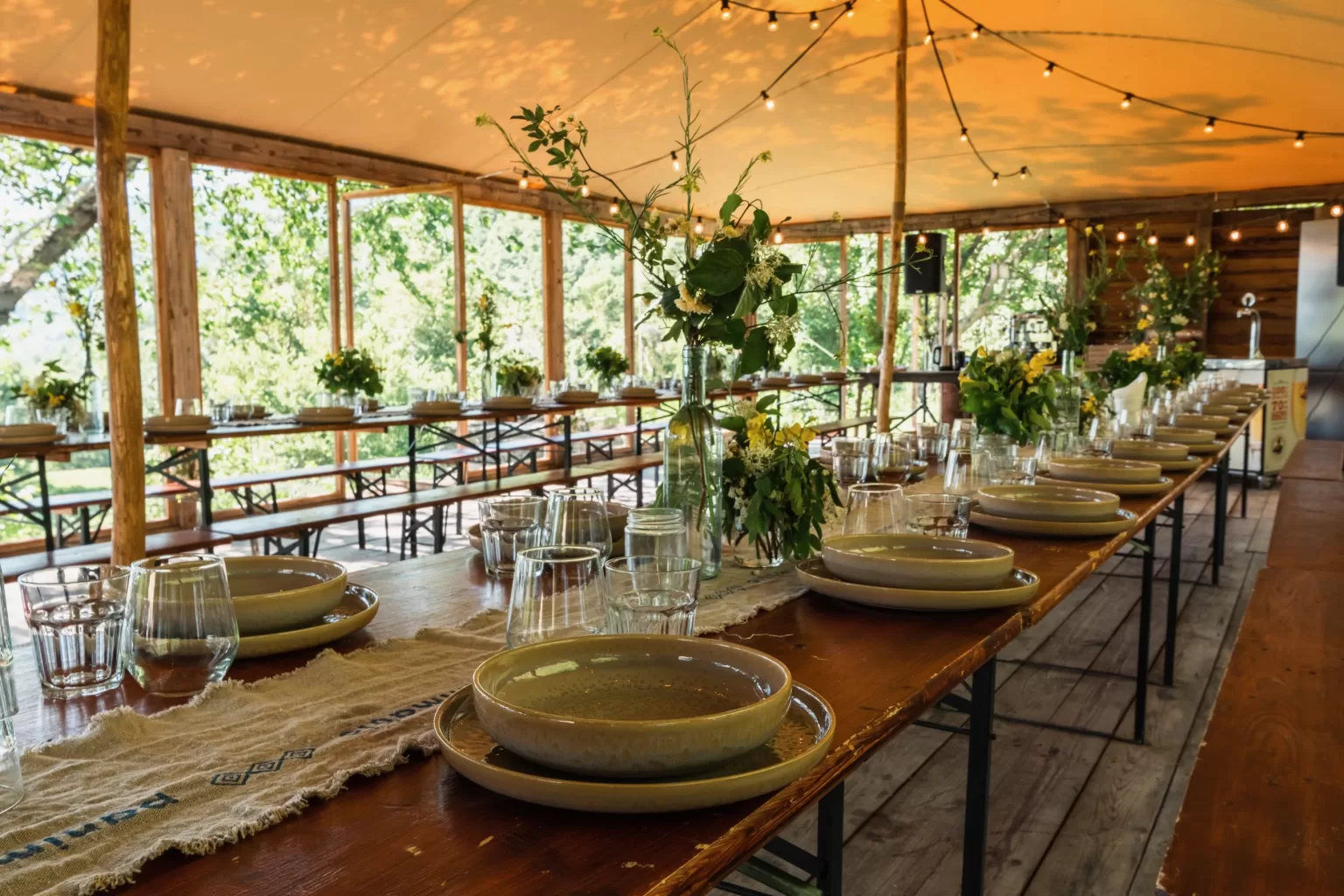 Long wooden tables set for a wedding reception under a tented terrace at Novanta 90 with natural light.
