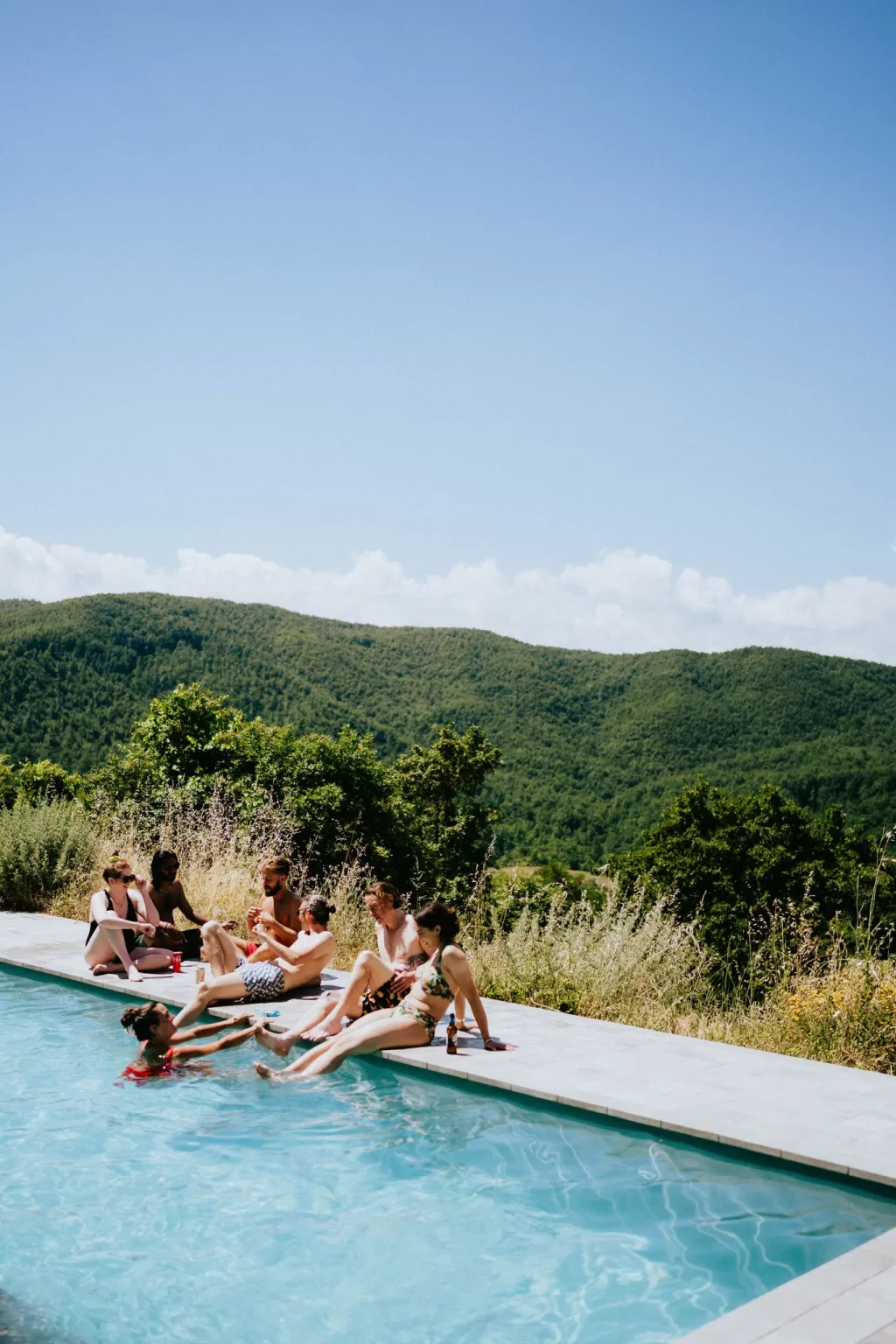 Friends relaxing by the pool at Novanta 90 on a sunny afternoon, surrounded by lush green hills.