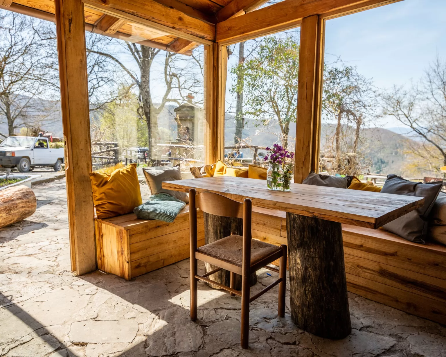 Wooden sunroom at Novanta 90 with large windows, cushioned benches, and mountain views bathed in golden light.