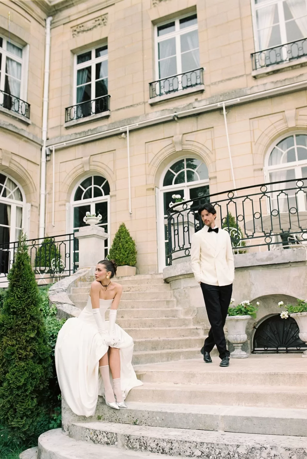 Bride seated on the lower steps while the groom stands on the balcony above at Château du Prieuré.