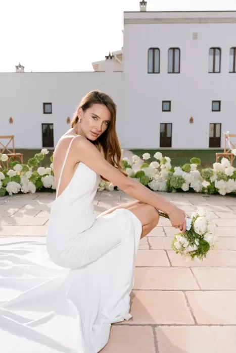 The bride kneeling outdoors at Apulien Bari in a sleek white gown, holding a bouquet with the villa behind her.