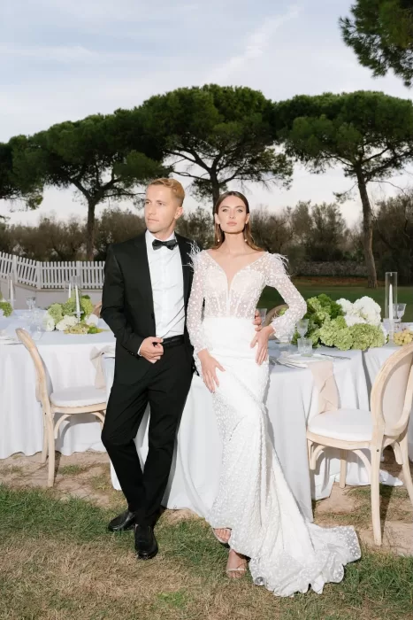 Bride and groom posing elegantly beside their outdoor reception setup surrounded by greenery in Apulien, Bari.