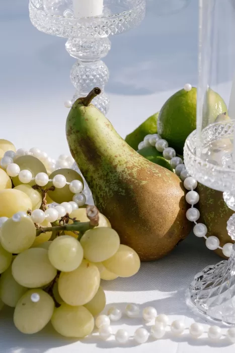 Still-life arrangement of pears, grapes, and limes with pearl strands at Apulien Bari, styled beside crystal candleholders.