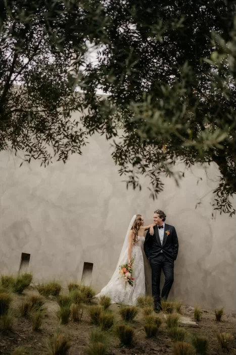 Bride and groom leaning together against a textured wall at Areias do Seixo, framed by olive trees.