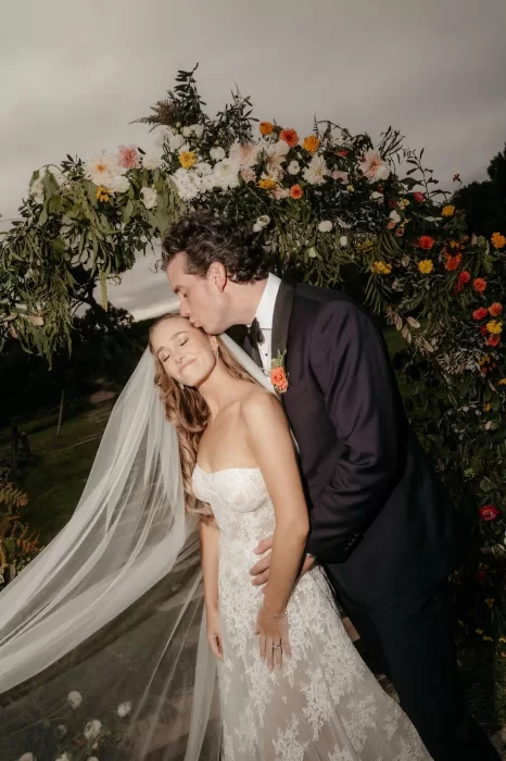 Groom kissing the bride beneath a lush floral ceremony arch at Areias do Seixo as her veil moves softly in the wind.