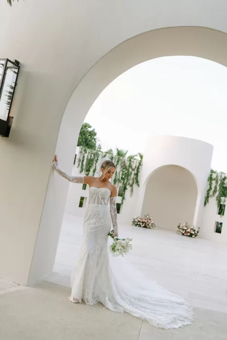 Bride standing beneath a white arch at Baja Luna, holding a bouquet and wearing a lace wedding gown.