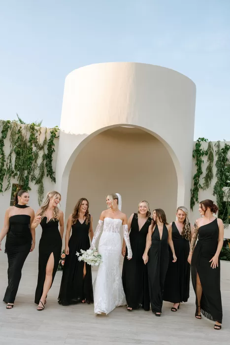 Bride walking with bridesmaids in black dresses beneath the sculptural ceremony arch at Baja Luna.