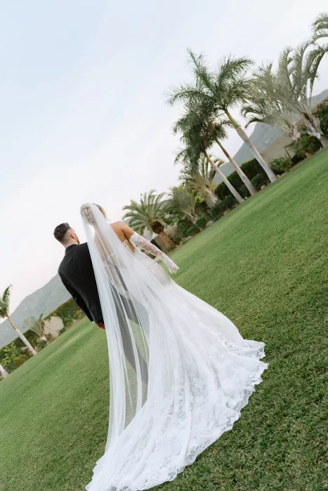 Bride and groom walking together across the lawn at Baja Luna with palm trees and mountains in the background.