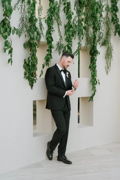 Groom in a black tuxedo standing beneath cascading greenery at Baja Luna before the ceremony.