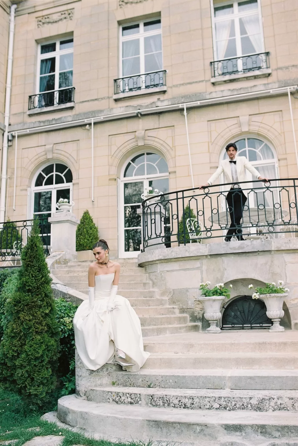 Bride seated on the lower steps while the groom stands on the balcony above at Château du Prieuré.