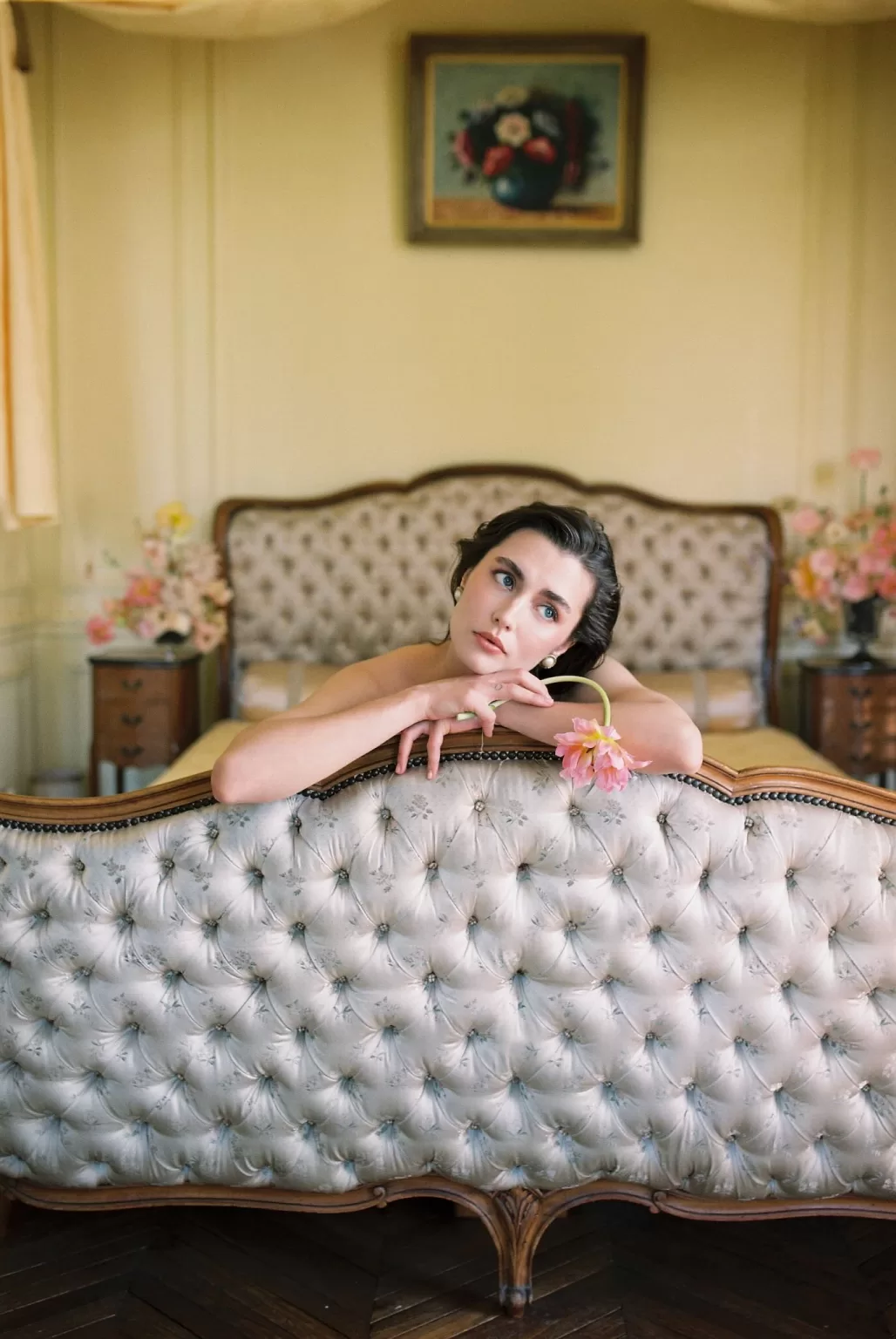Bride resting her arms on an ornate tufted bedframe in a serene bedroom at Château du Prieuré, holding a delicate flower in a dreamy moment.