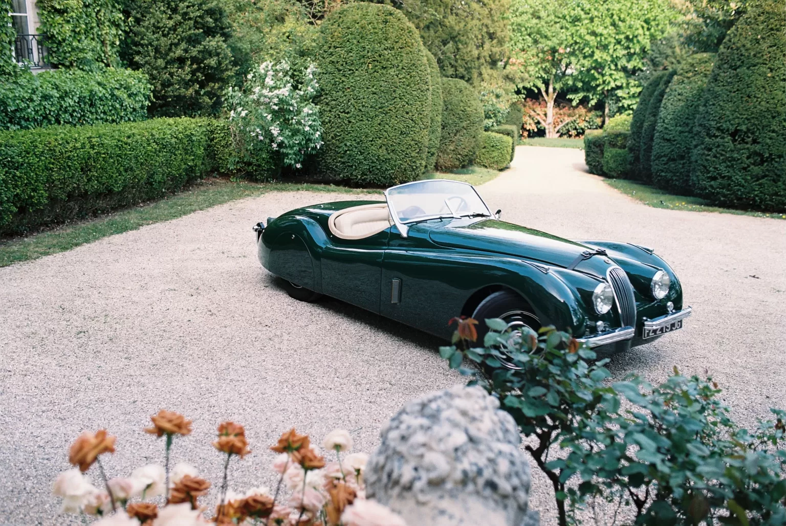 A classic green roadster parked in the garden courtyard of Château du Prieuré, framed by manicured hedges and romantic blooms.