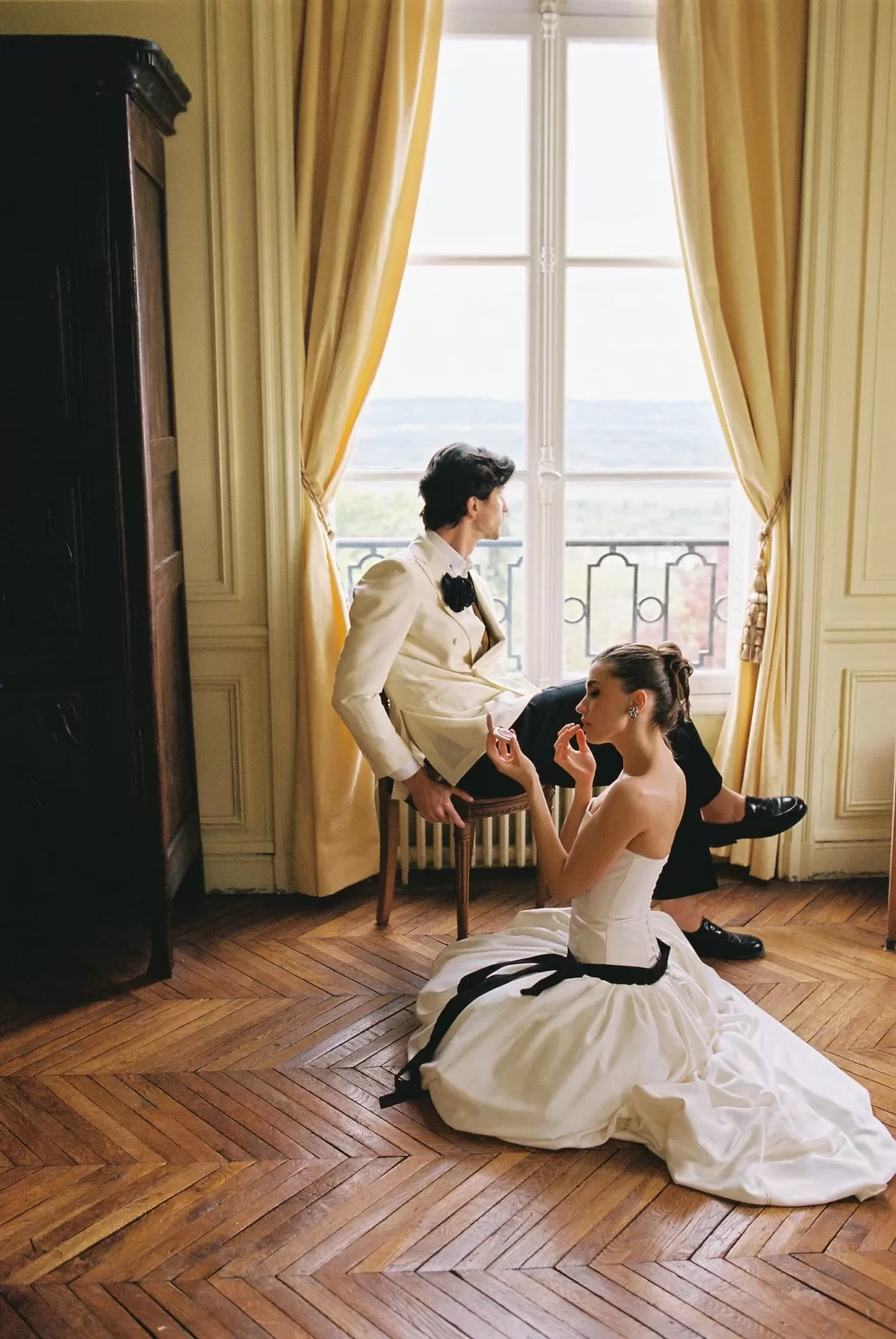 Bride and groom in an elegant room at Château du Prieuré, framed by tall golden drapes and a view of the countryside.