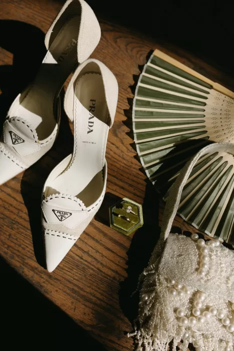 Bridal accessories flat lay featuring Prada heels, wedding rings, pearl handbag and folding fans styled at Chateau Lill.
