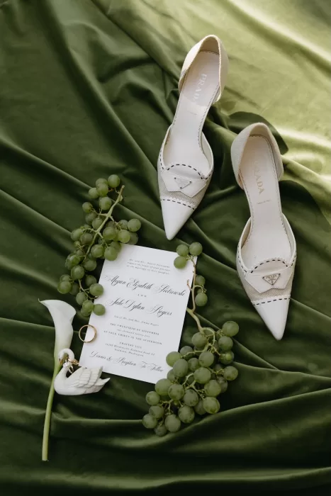 Bridal flat lay featuring Prada shoes, wedding invitation and green grapes styled on silk fabric at Chateau Lill.