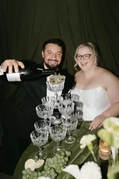 Bride and groom laugh as they pour champagne into a crystal glass tower at Chateau Lill, styled with green velvet linens and grapes.