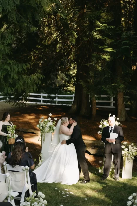 Bride and groom share their first kiss during a woodland ceremony at Chateau Lill, framed by towering trees and white floral arrangements.